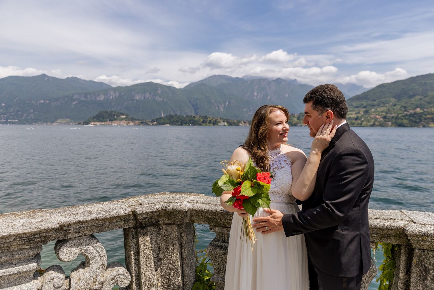 Newlyweds embracing at lakeside terrace with mountain backdrop and colorful bouquet.