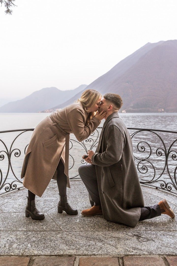 Couple sharing a kiss during a marriage proposal on a lakeside terrace with mountain backdrop.