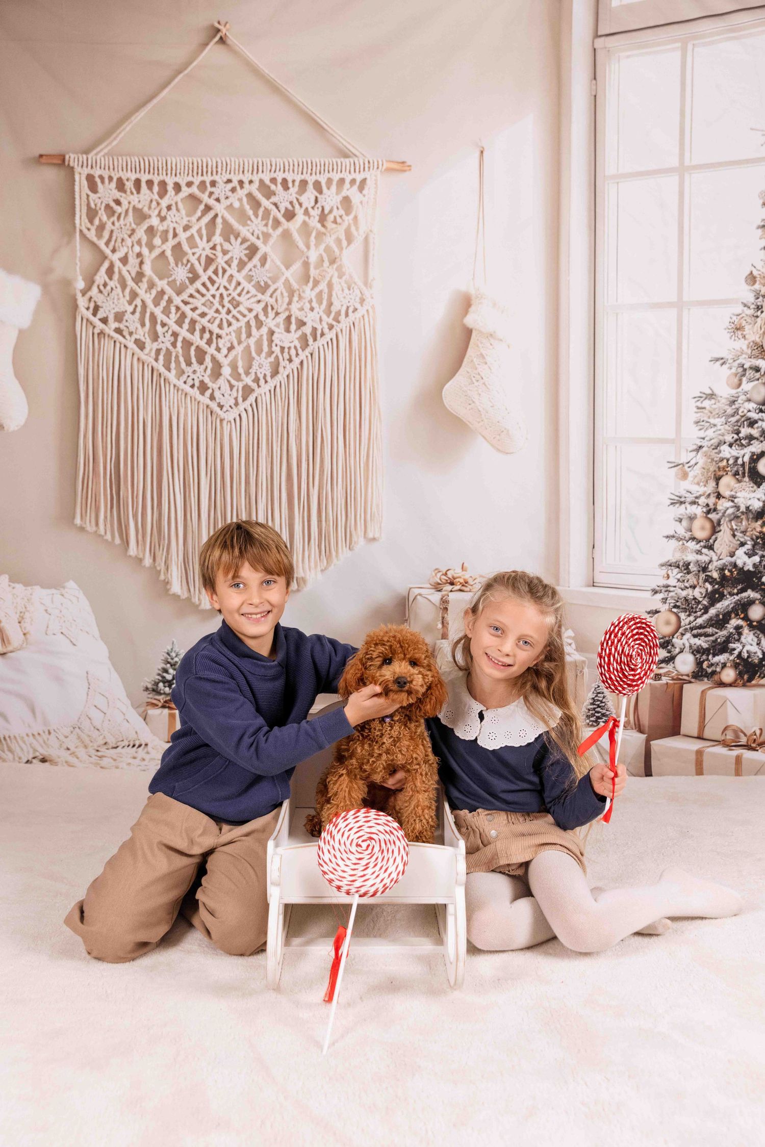 Two children and brown dog pose on floor with candy canes near decorated Christmas tree and macrame wall art.