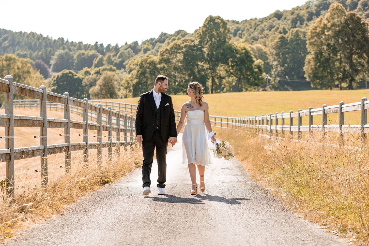 Newlyweds walk along rustic fence-lined path through golden fields.