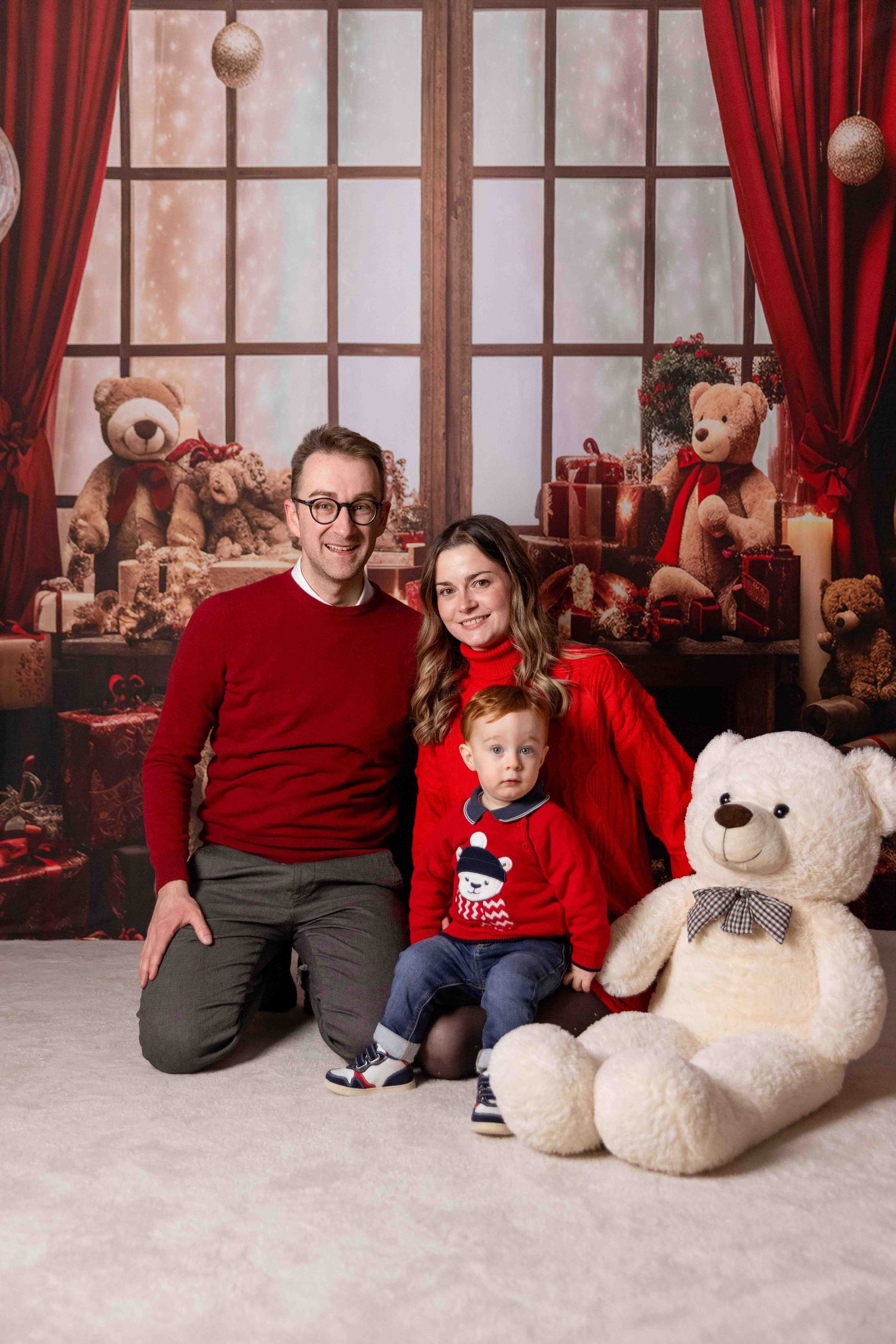 Cozy Christmas scene with red curtains and plush teddy bears on snowy white floor.