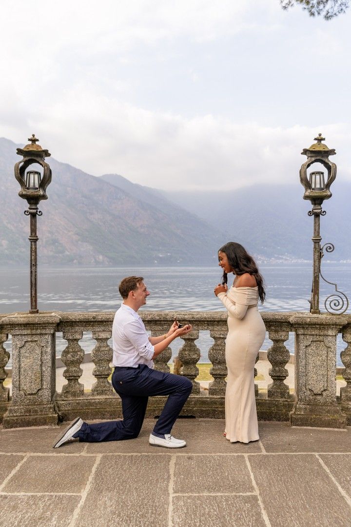 Man proposes on one knee to woman in white dress on scenic lakeside terrace with mountain backdrop.