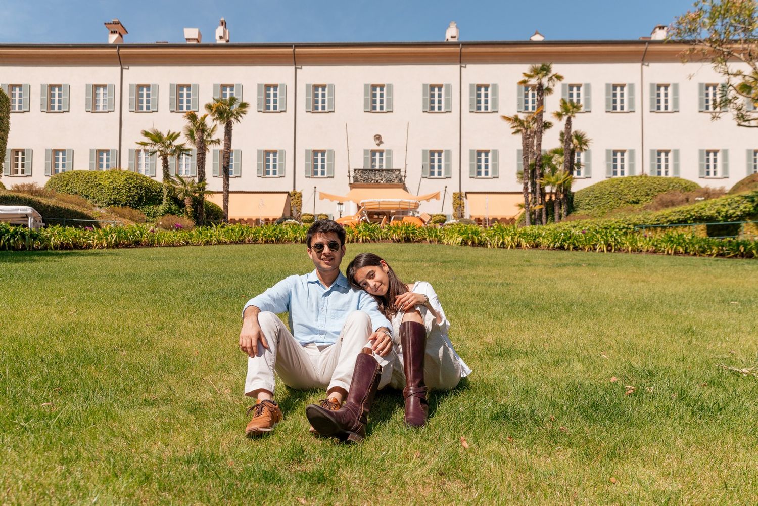 Couple sitting on green lawn in front of elegant white Italian villa with palm trees on sunny day.