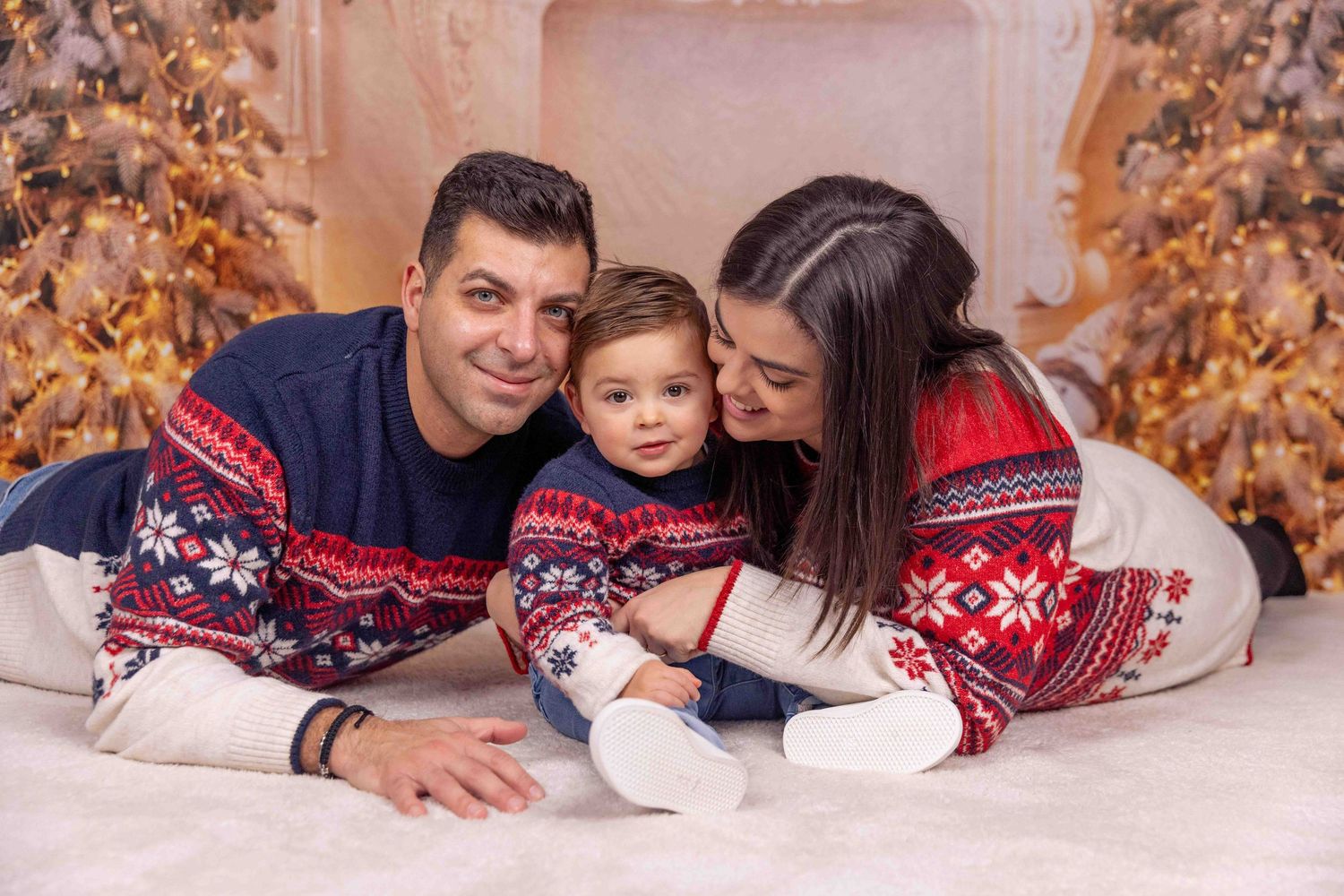 Family wearing matching red and navy Nordic sweaters poses together on white floor during holiday photoshoot.