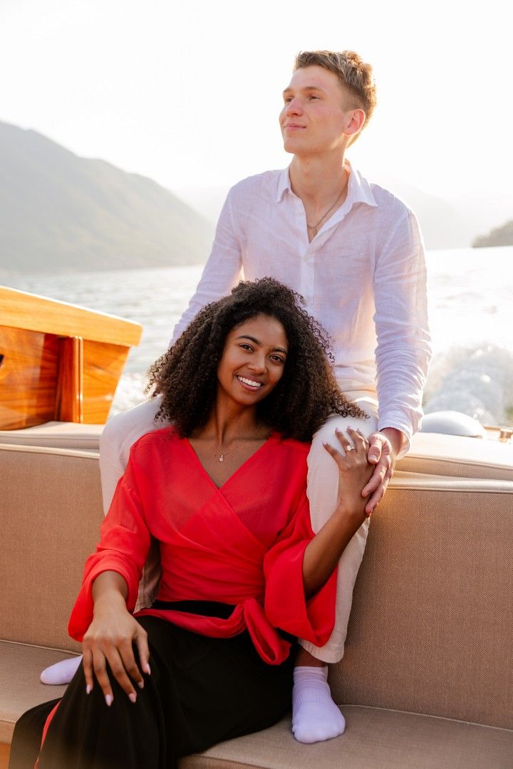 A smiling interracial couple holding hands on a yacht, woman in red top, man in white shirt, mountains in background.