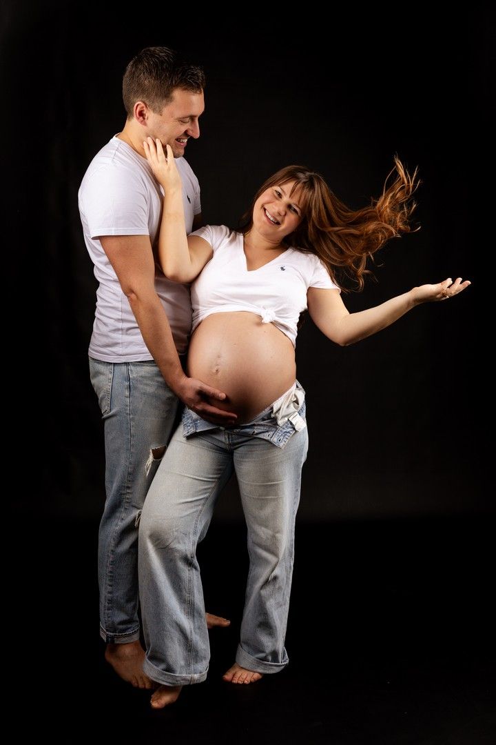 Happy pregnant couple posing together, man holding woman's baby bump, both smiling, wearing jeans and white shirts.