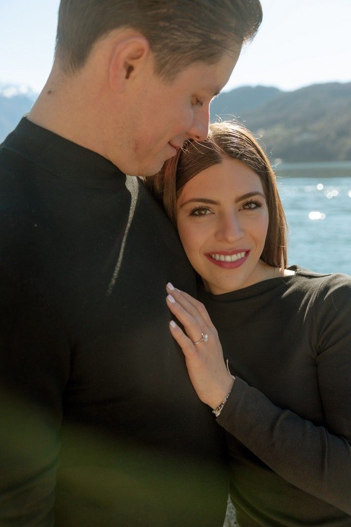 Couple embracing outdoors near a lake with mountains, woman smiling and showing engagement ring.