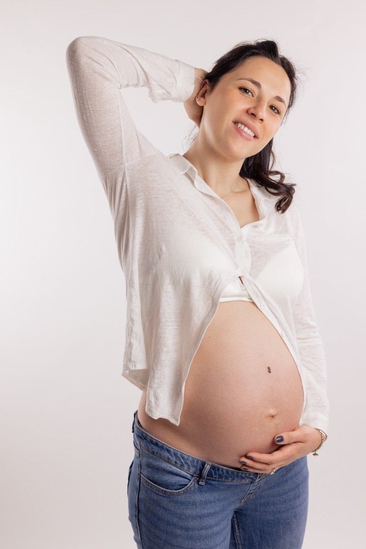 Casual maternity portrait of woman in white shirt and jeans against white backdrop.