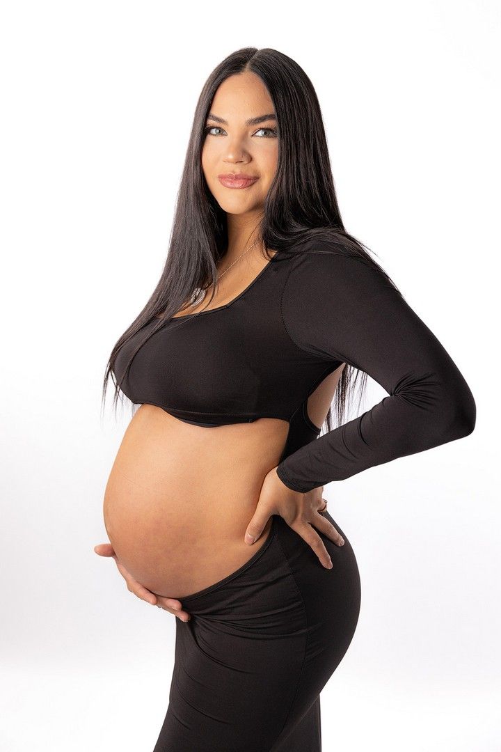 Side view of pregnant woman in black outfit showing her baby bump against white backdrop.