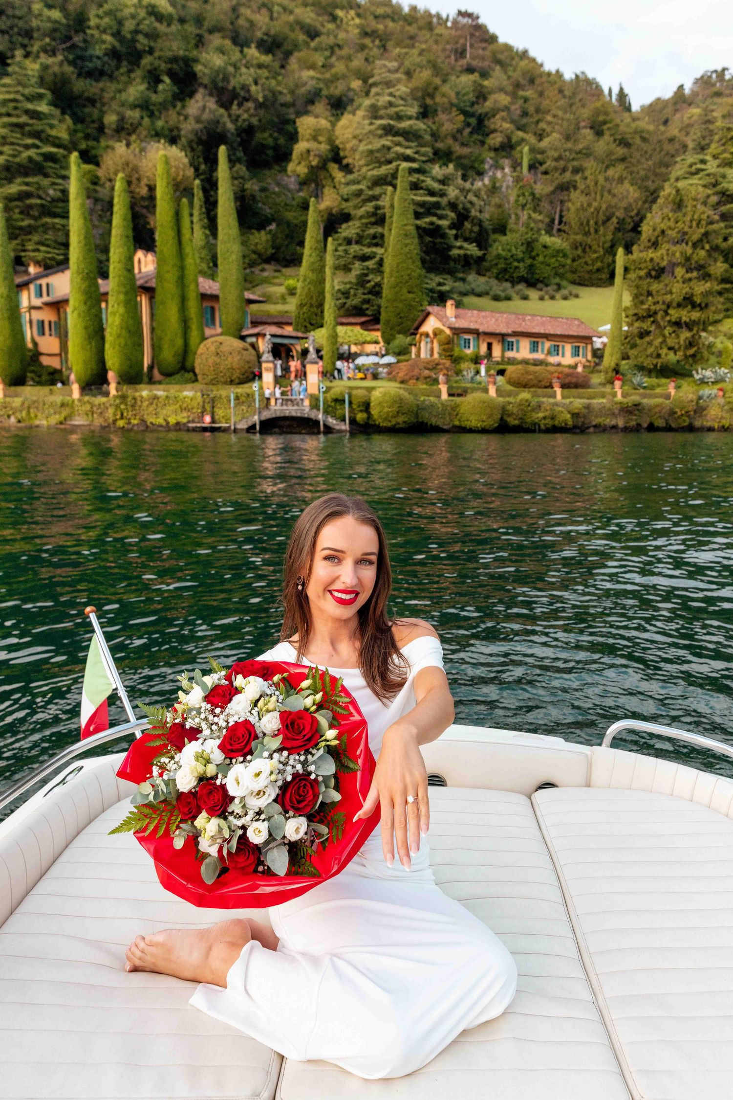 A scenic boat photoshoot on Lake Como featuring red roses and cypress trees in the background.