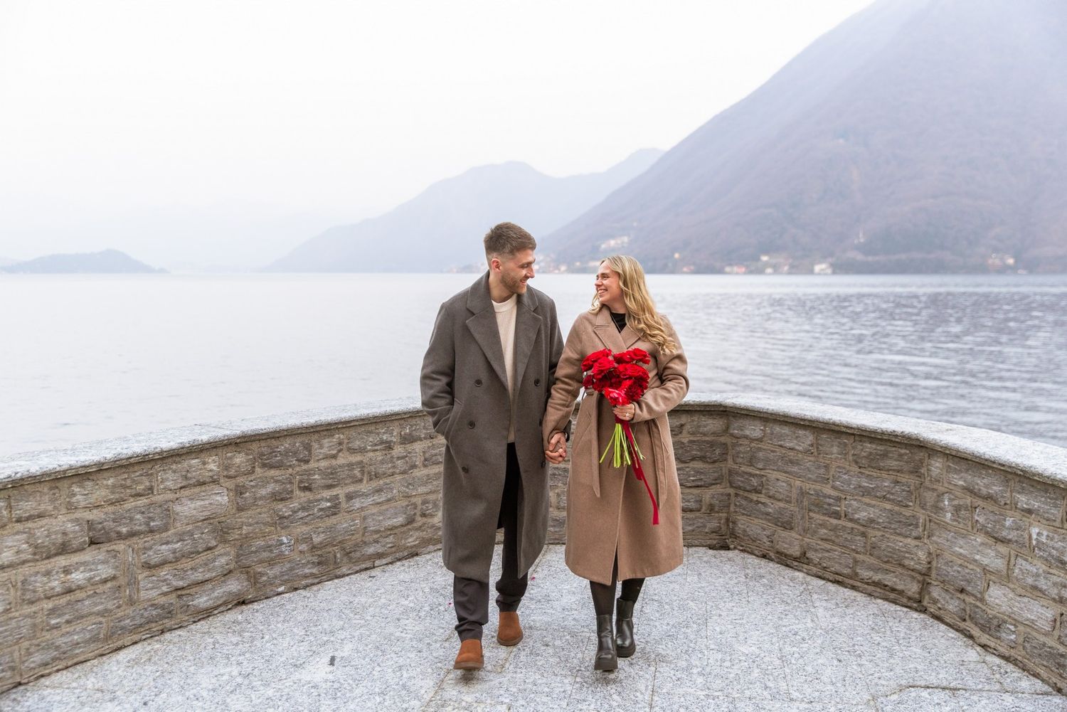 Couple walking on a lakeside terrace in winter, woman holding red roses, mountains in the misty background.