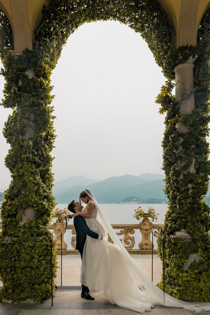 Wedding couple shares romantic moment under ivy-covered arches overlooking lake.