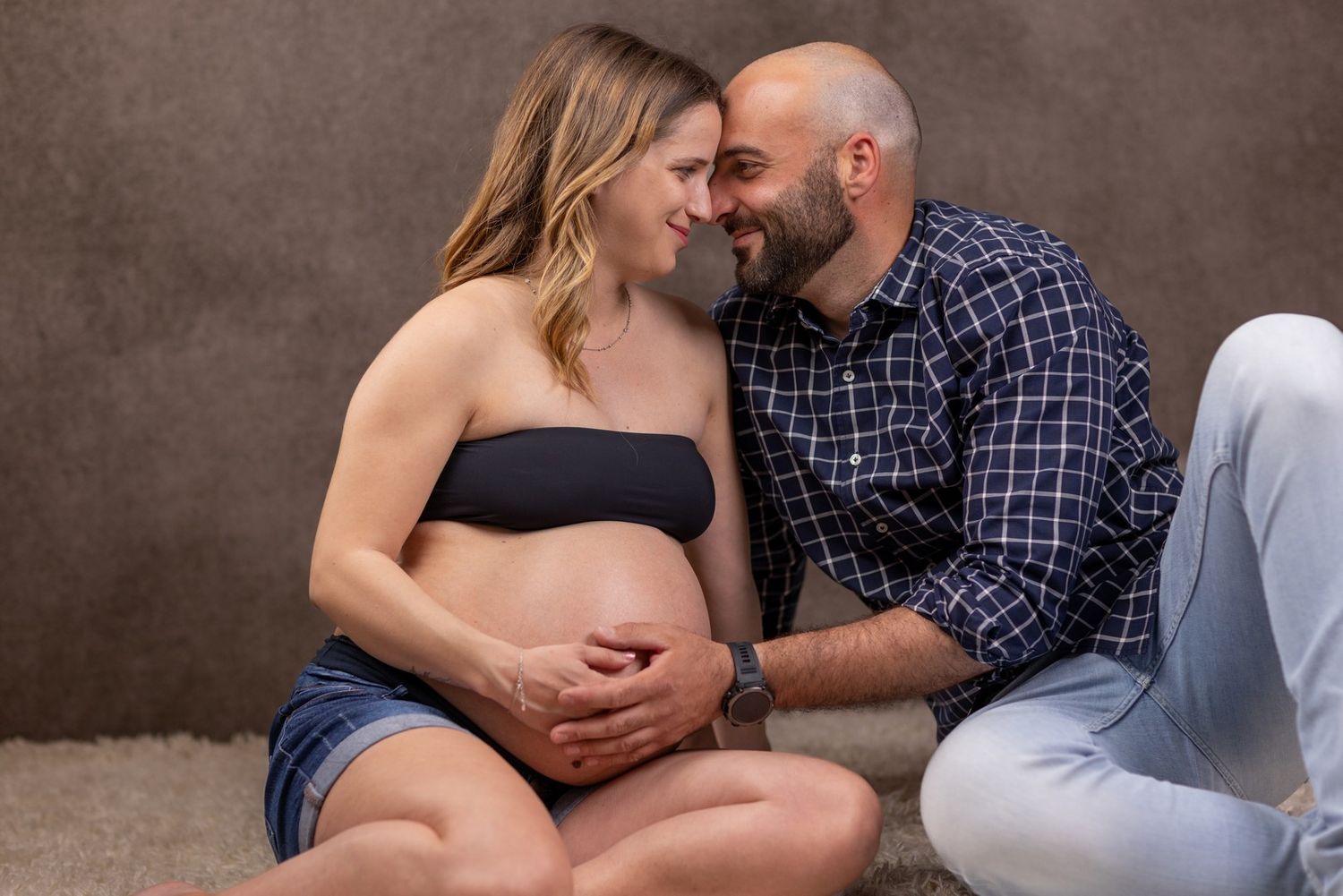Intimate maternity photo of couple sitting together against brown backdrop.