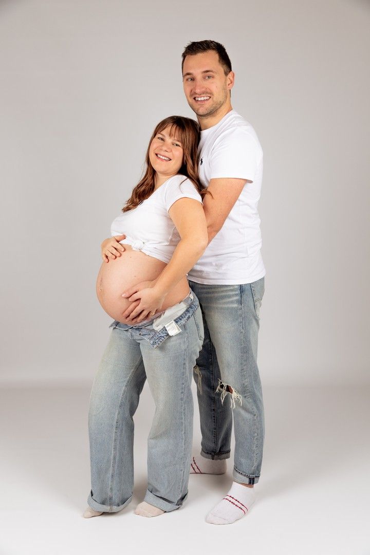 Happy couple in matching white tees and jeans, man embracing pregnant woman showing baby bump in studio.