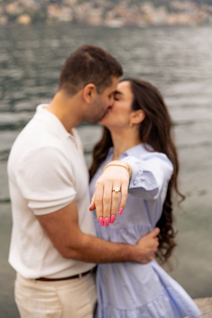 Couple kissing while woman shows off engagement ring by a lake.