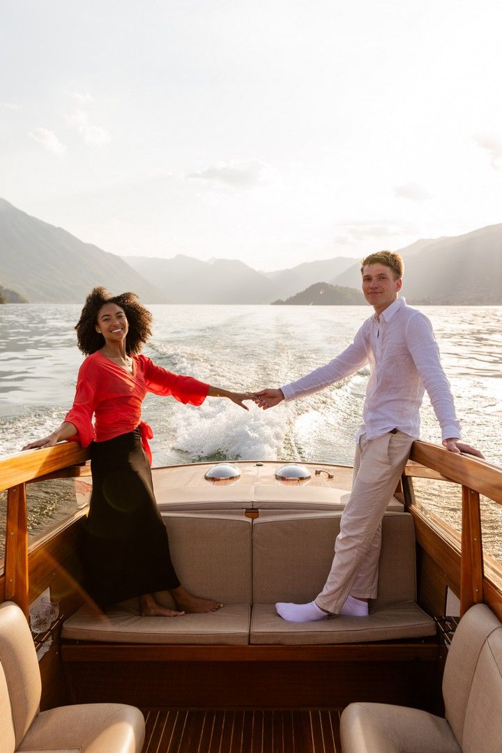 Couple holding hands on a luxury boat with mountain lake backdrop at sunset, woman in red top, man in white shirt.
