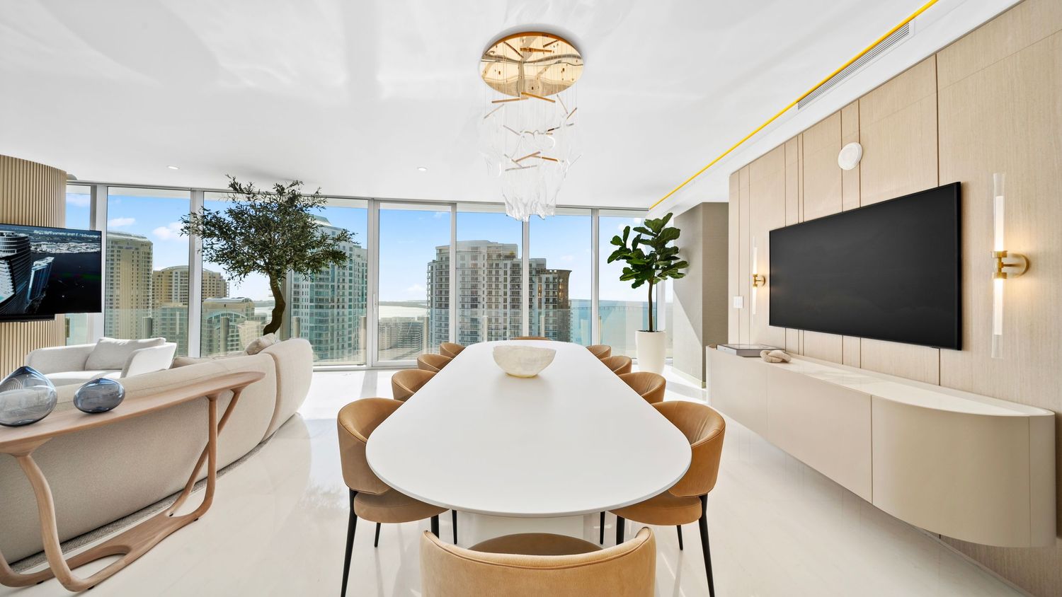 Modern dining room with white oval table, tan chairs, wall-mounted TV, and panoramic city views through floor-to-ceiling windows.