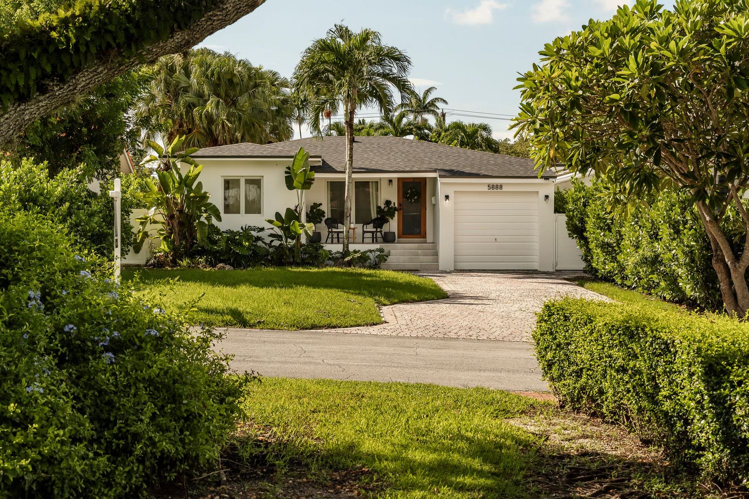 Modern white ranch house with palm trees and lush landscaping features curved driveway and attached garage.