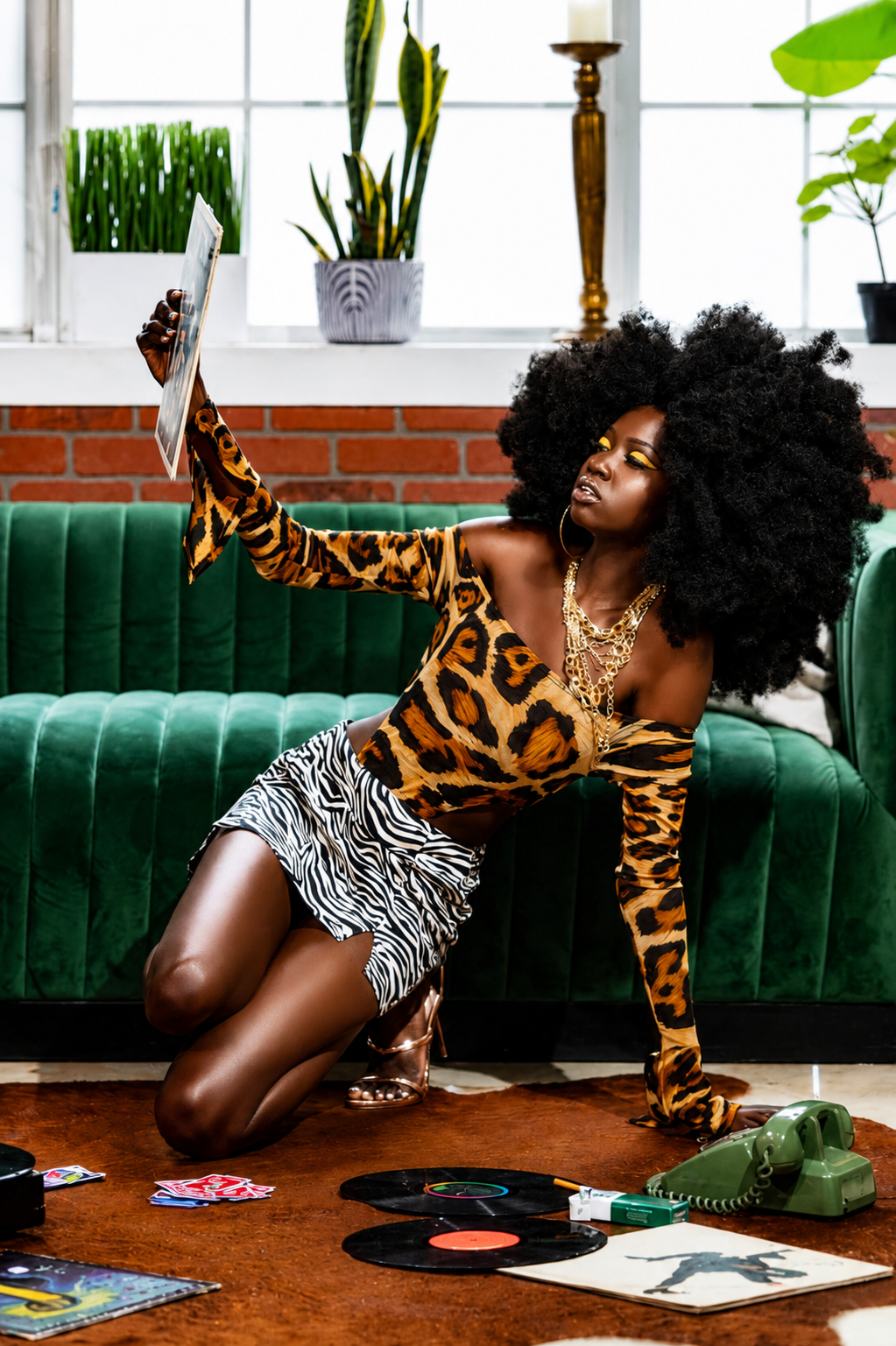Woman in leopard print outfit with afro hairstyle poses on floor with vinyl records near green velvet sofa.