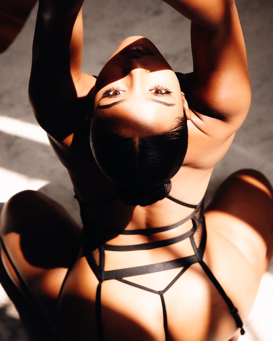 Woman in black strappy lingerie posing in dramatic shadows and sunlight, looking upward with arms raised.
