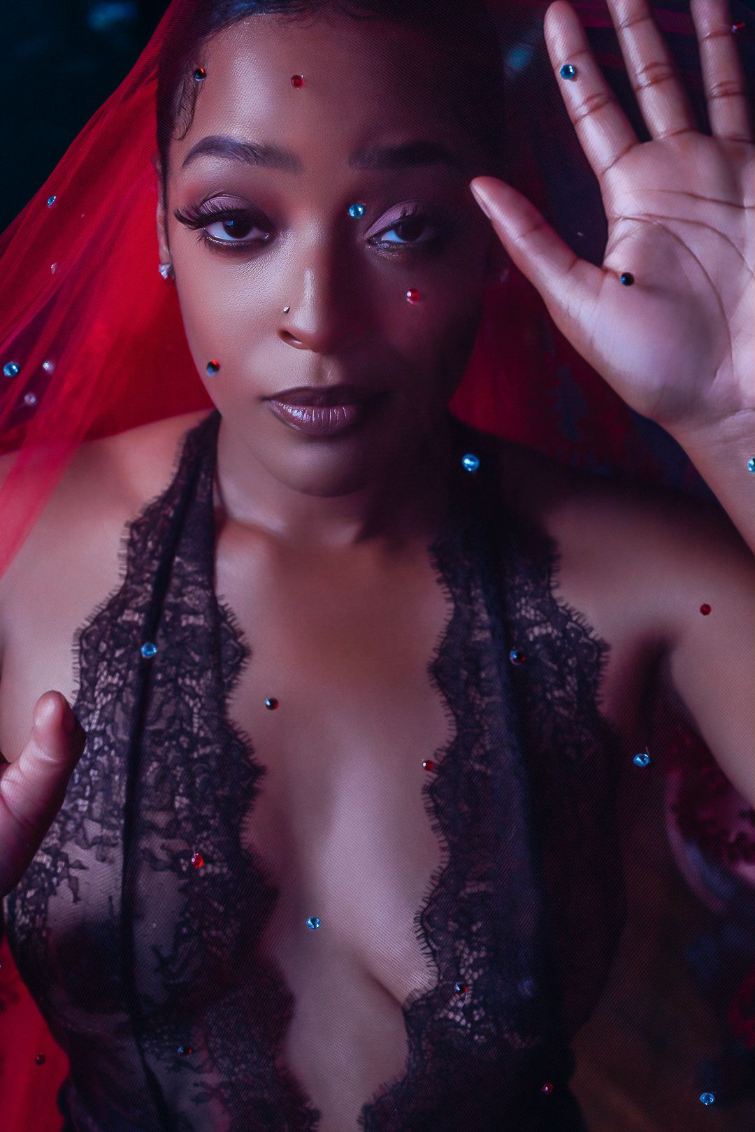 A woman adorned with colorful gemstones and a red veil poses in black lace lingerie under dramatic lighting.