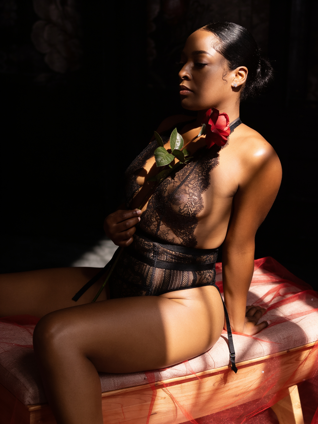A woman in black lace lingerie holds a red rose while posing gracefully in dramatic studio lighting.