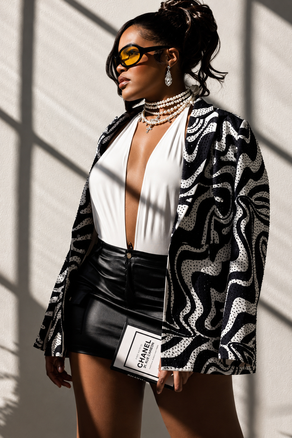 Woman in patterned sequin blazer, white bodysuit, leather skirt, and yellow sunglasses holds Chanel book.
