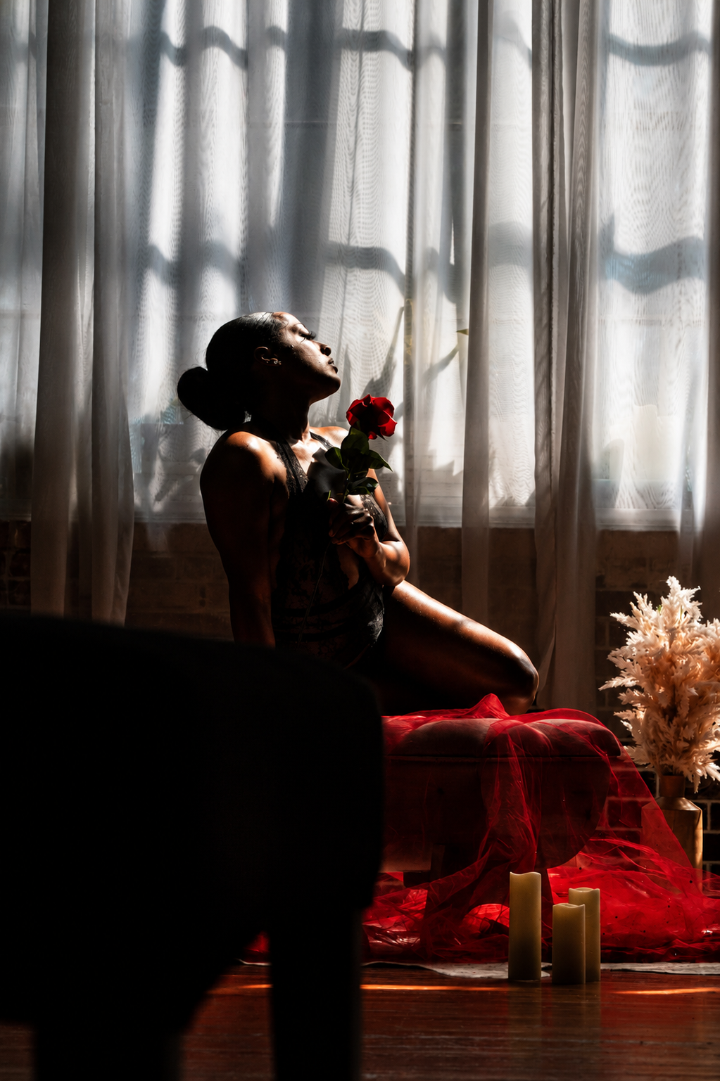 A woman sits gracefully holding a red rose, silhouetted against sheer curtains with candles nearby.