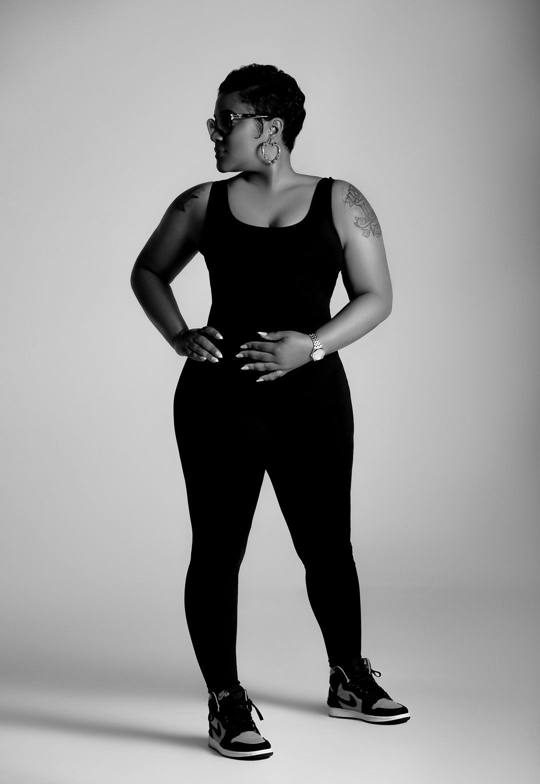 Black and white portrait of a woman in all-black outfit and sneakers posing confidently in a studio.