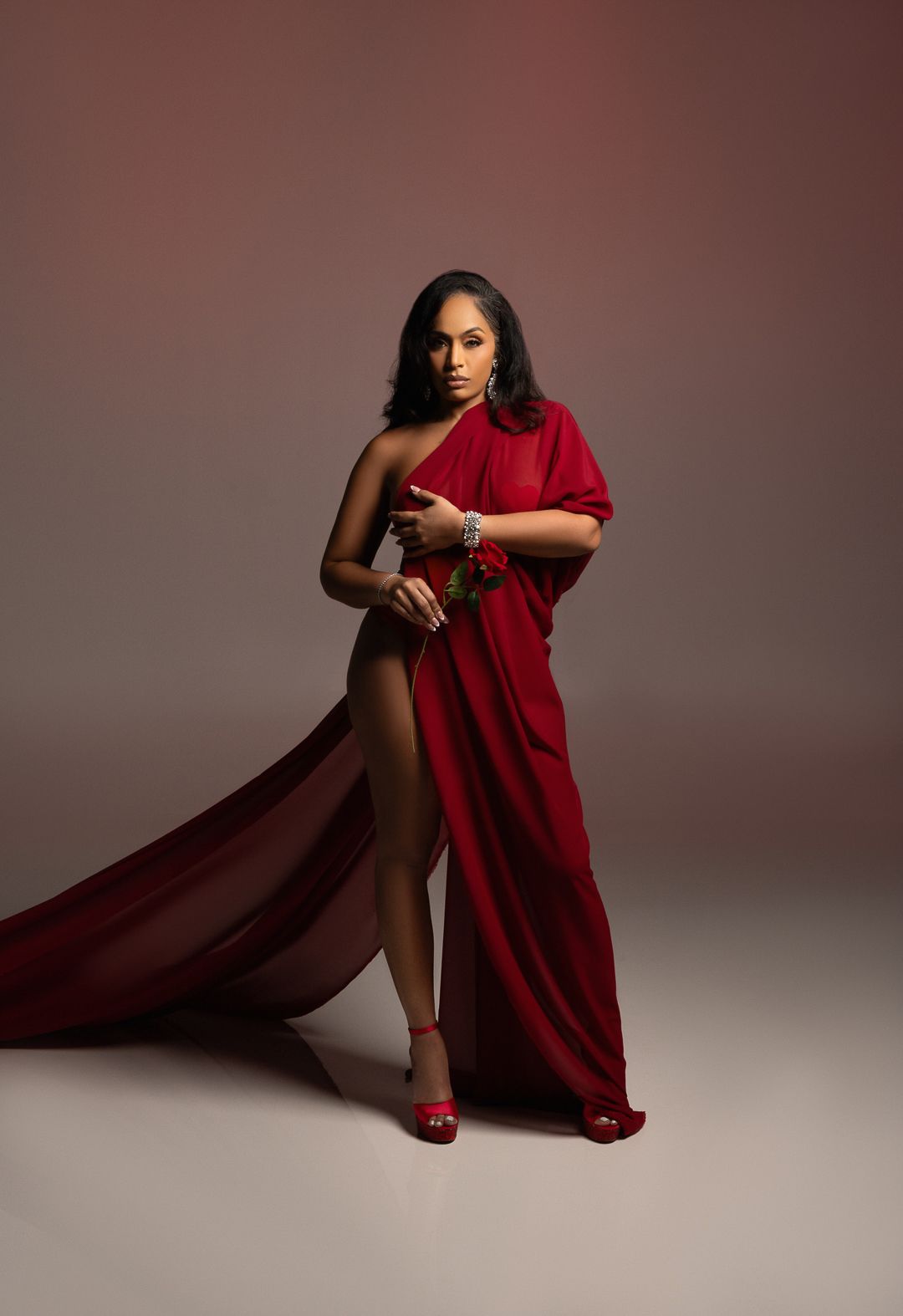 Woman in elegant red one-shoulder gown with flowing train and matching heels poses gracefully in studio.