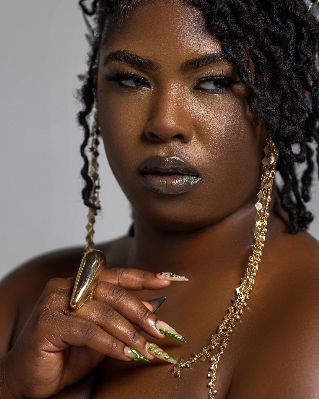 A Black woman with locs, metallic lips, gold nails, and layered gold jewelry poses confidently against a gray background.
