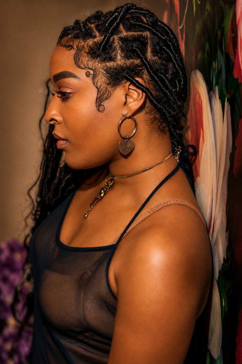 A Black woman with braided locs, hoop earrings, and a gold necklace wears a sheer black top, posing sideways near floral art.