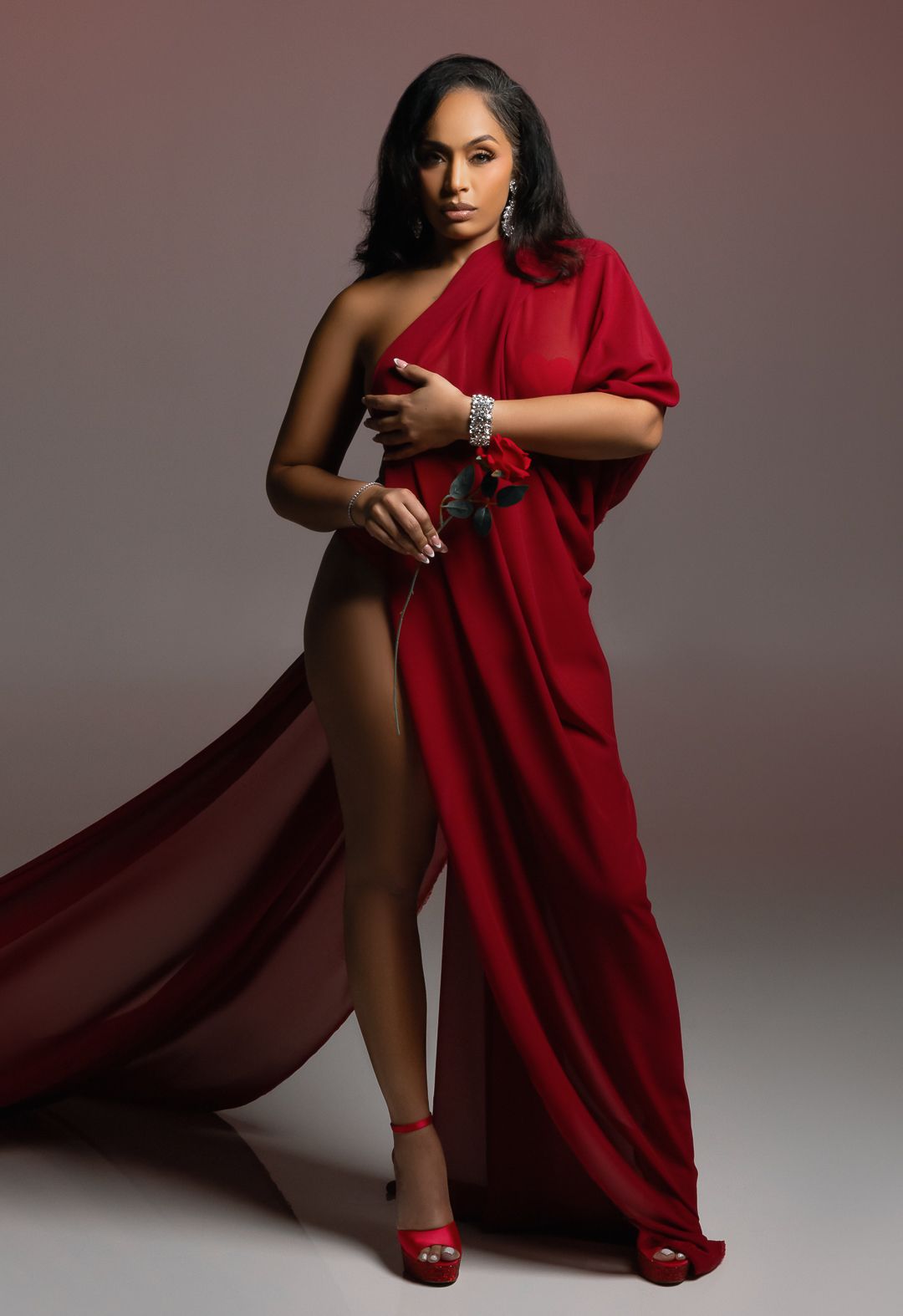 Woman in elegant red one-shoulder flowing gown with high slit, wearing silver jewelry and red heels, posing gracefully.
