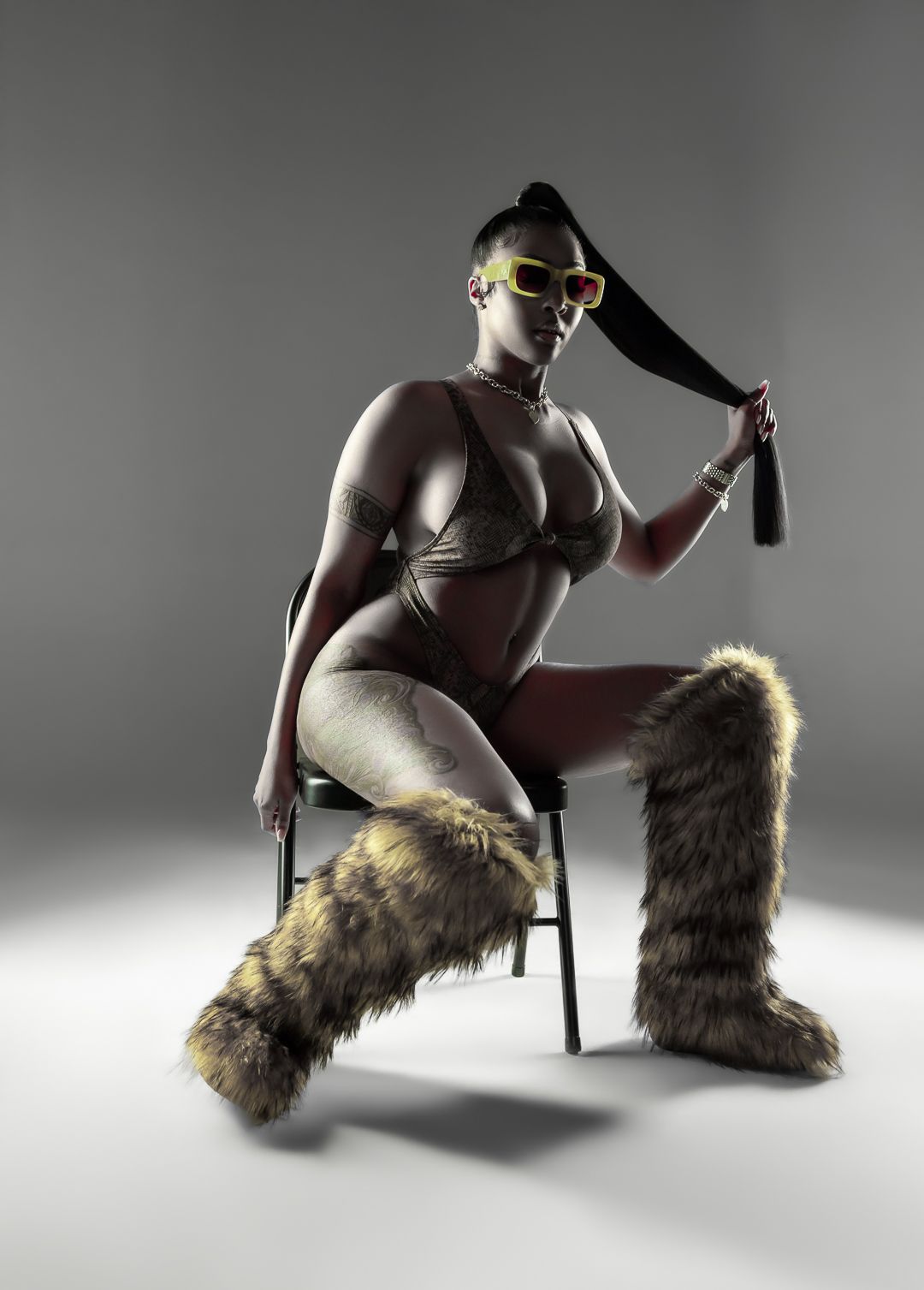 A woman poses in a bikini with faux fur boots, yellow sunglasses, and a long ponytail in a studio setting.