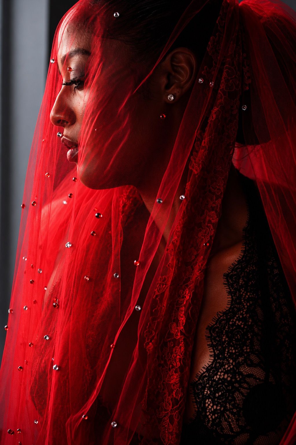 Woman in black lace wearing a red jeweled veil, profile view in dramatic lighting.