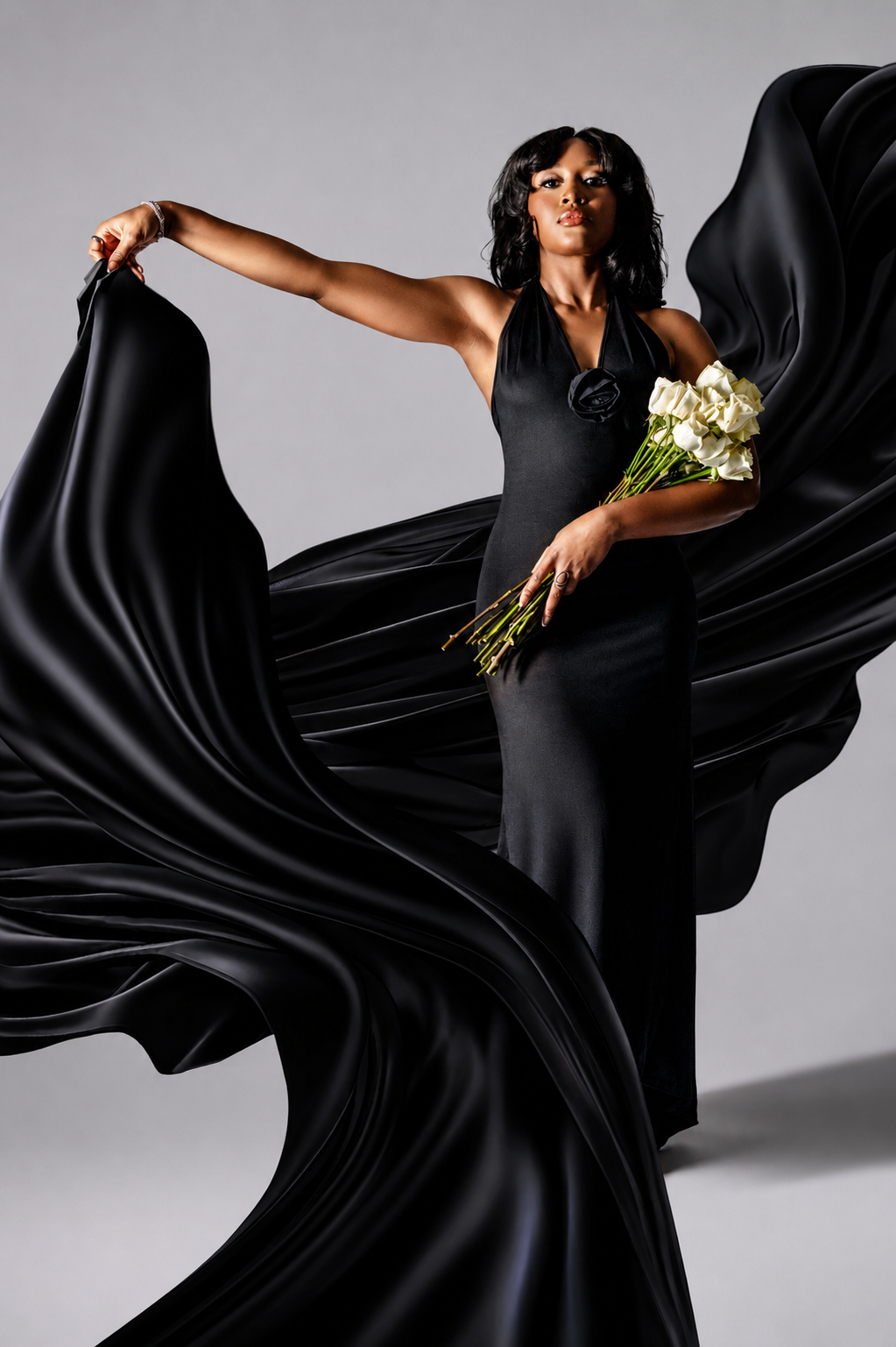 Woman in elegant black satin gown with flowing fabric holds white roses in a dramatic studio pose.