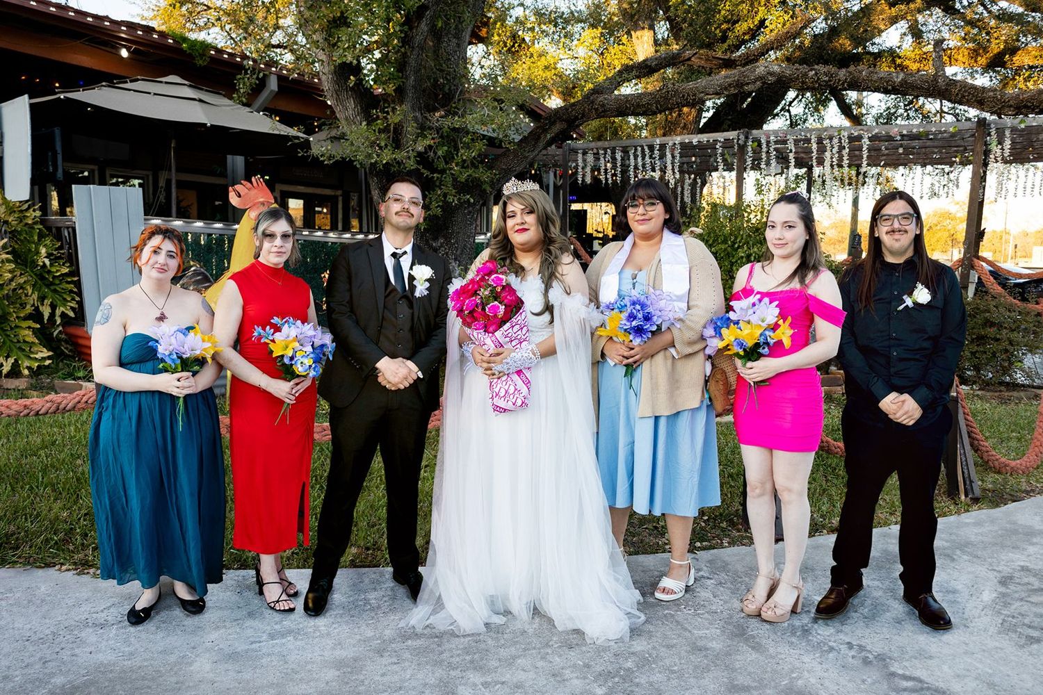 Wedding party posing outdoors with bride in white gown, groom in tuxedo, and colorful bridesmaid dresses.