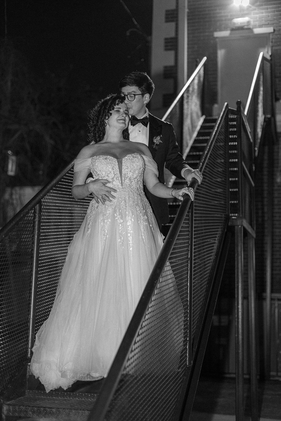 A couple in formal attire share a romantic moment on a staircase at night with dramatic lighting and shadows.