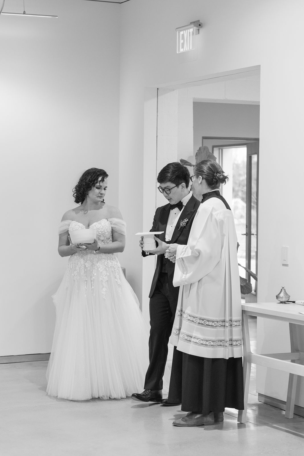 A black and white wedding ceremony photo showing a bride in a ballgown with an officiant and groom in formal attire.