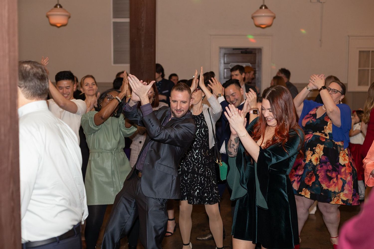 Wedding guests pose together with hands raised in a synchronized dance move at an indoor reception celebration.
