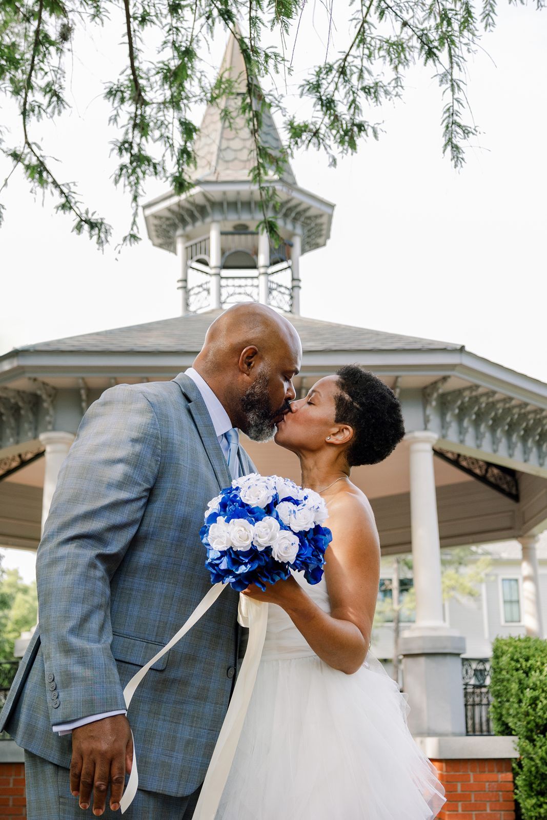 A romantic wedding couple shares intimate kisses in front of a white gazebo while holding a blue and white bouquet.