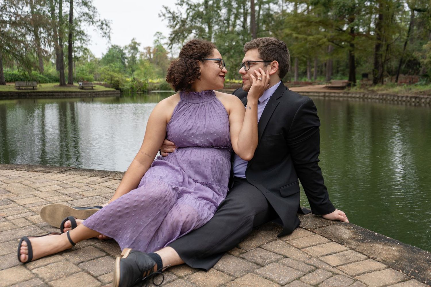 Couple sitting by a pond in a park, woman in purple dress and man in black suit sharing a romantic moment.