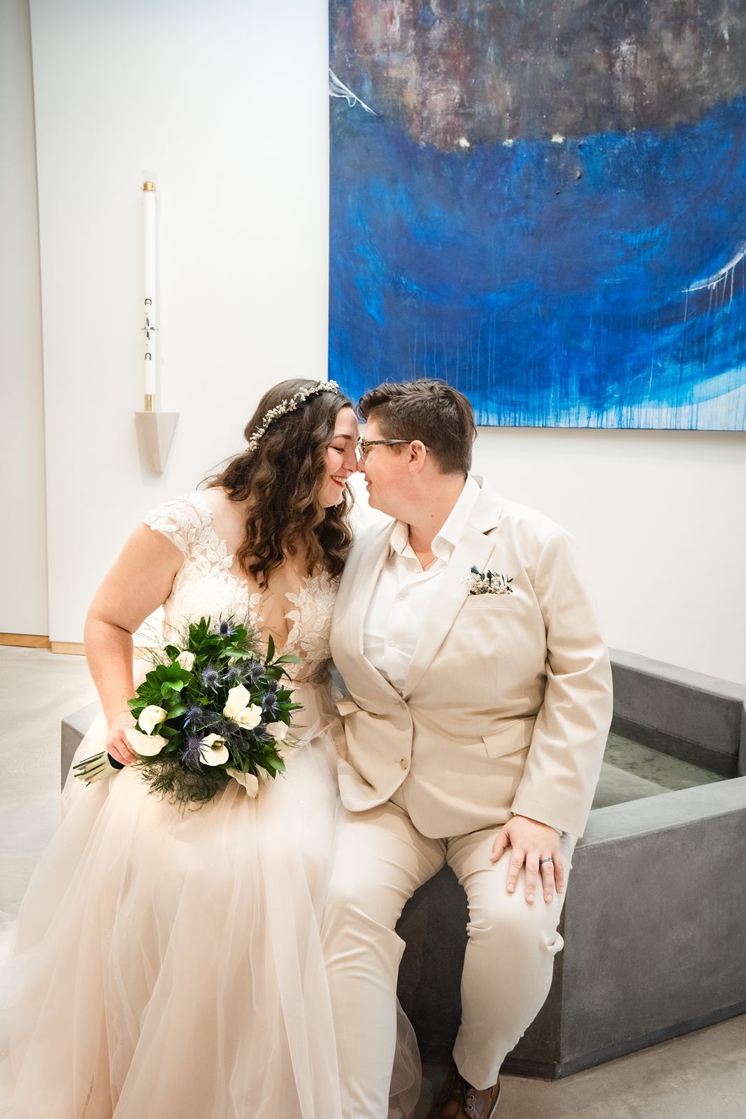 Two brides share a tender moment in front of a blue abstract painting, one in a white gown holding a bouquet.