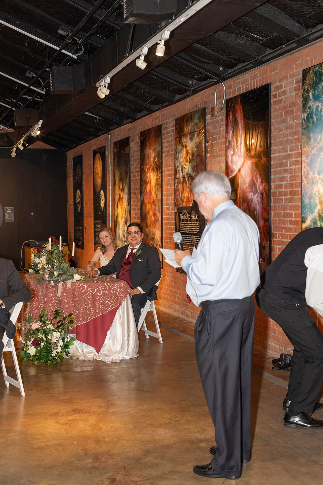 Guest delivers speech at wedding reception while couple sits at decorated sweetheart table.