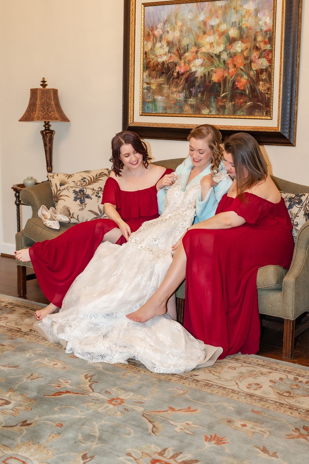 Bride in white lace gown gets help from two bridesmaids in red dresses while sitting on a sofa.