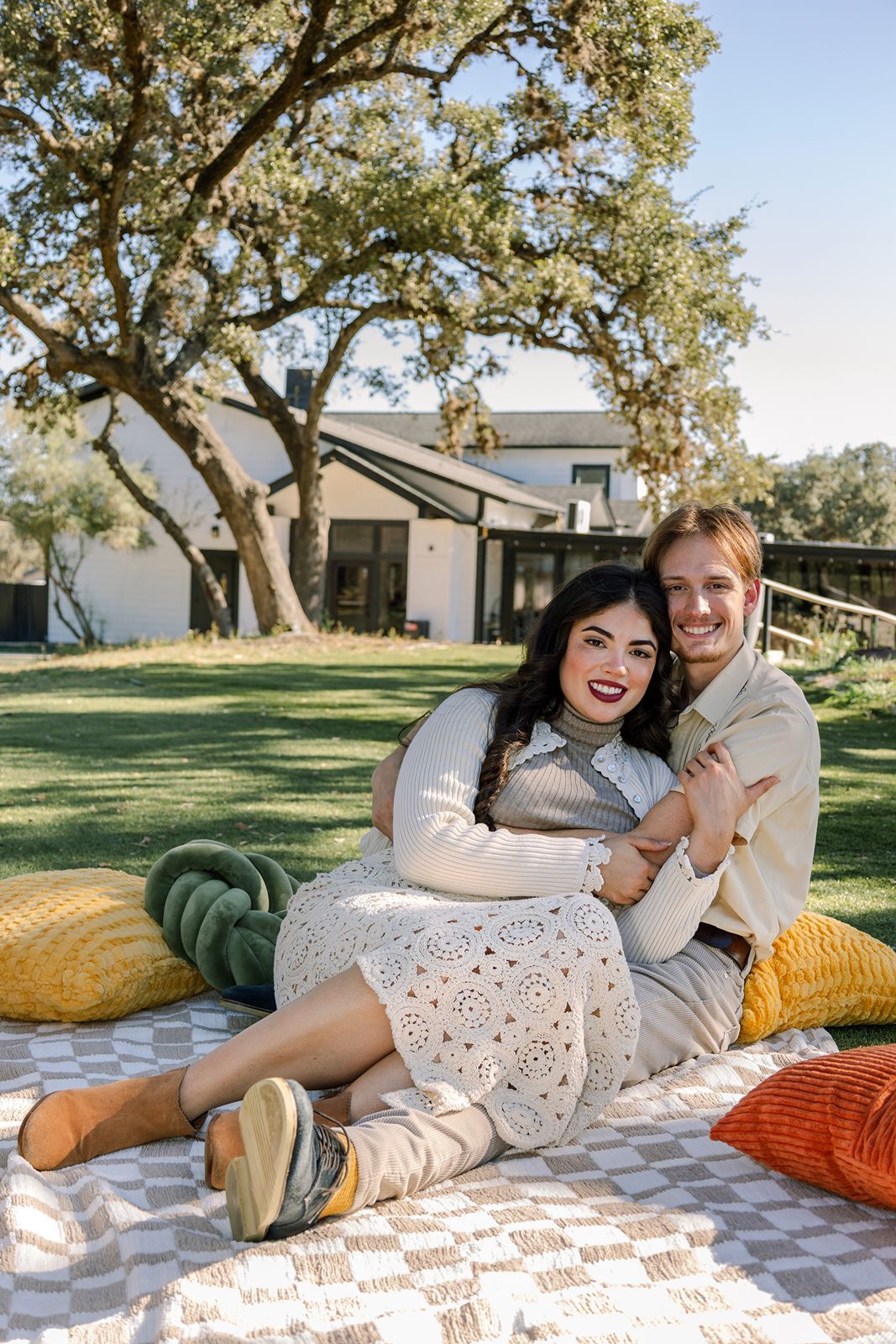 Couple enjoys romantic picnic on blanket with colorful pillows under large oak tree at modern house.