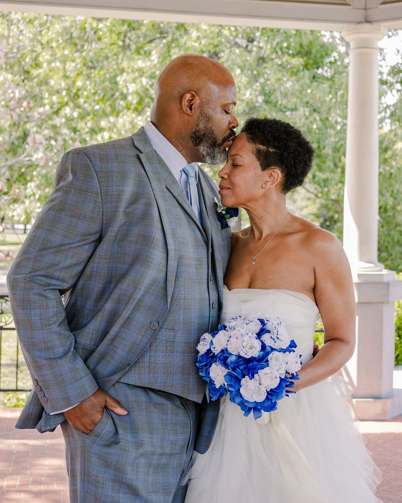 Bride and groom share intimate moment under gazebo, bride holding blue and white floral bouquet in elegant white dress.