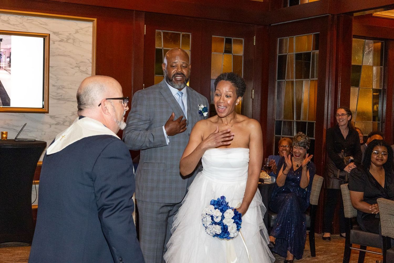 Bride in white gown with blue bouquet and groom in suit share a joyful wedding ceremony moment with guests.
