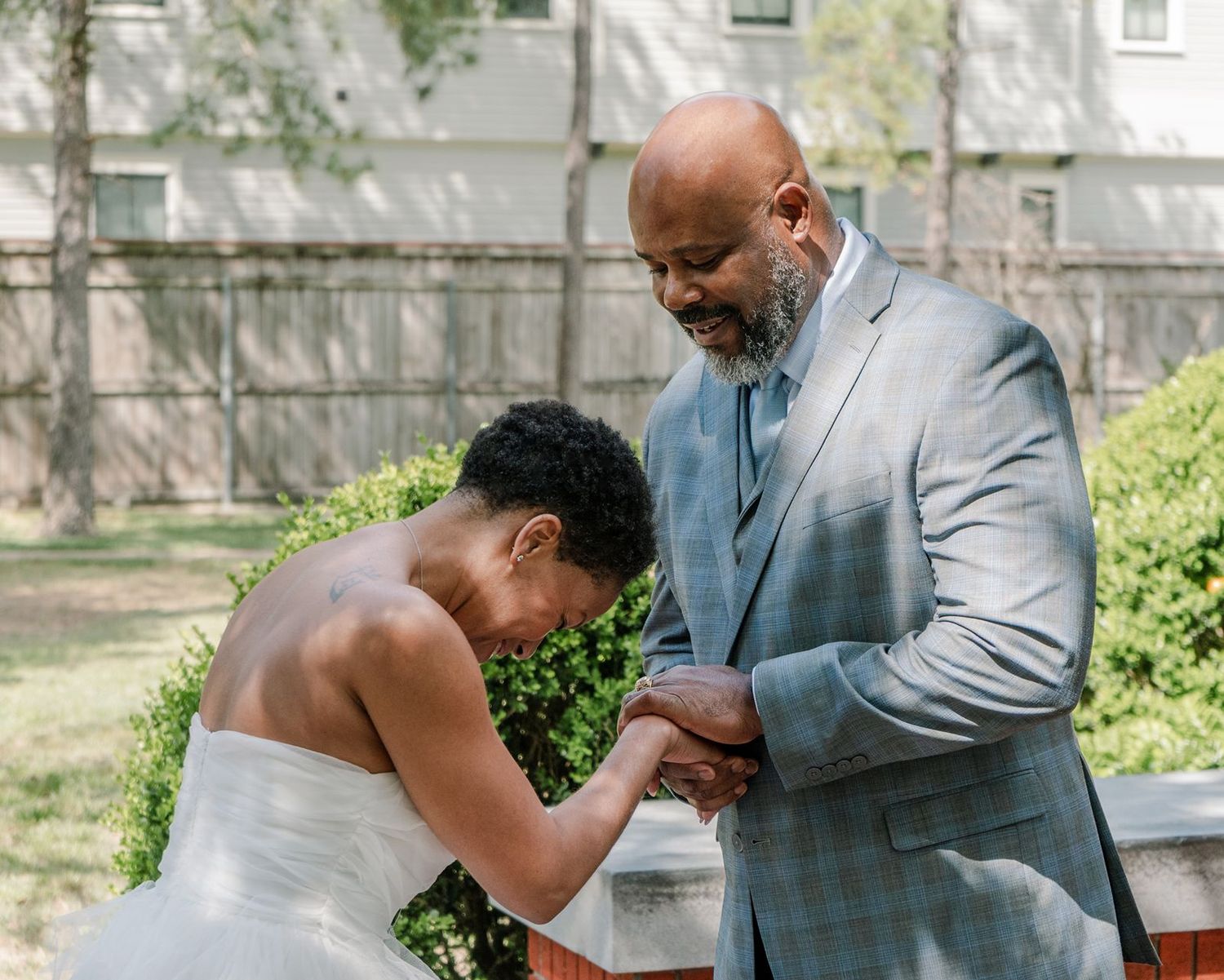 A bride in a white gown bows her head as the groom holds her hands tenderly outdoors.