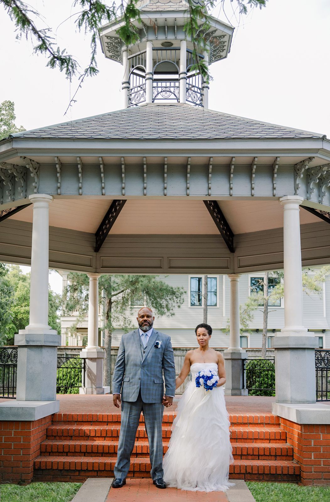 Bride in white gown and groom in gray suit posing formally in front of a classic white gazebo.